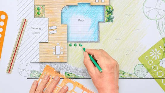 Skizze eines Gartens. Darin eingezeichnet eine große Holterrasse und ein Pool sowie Rasenfläche. Auf der Skizze liegen mehrere Zeichenutensilien. Eine Hand hält einen grünen Stift und zeichnet Büsche auf der Skizze ein.